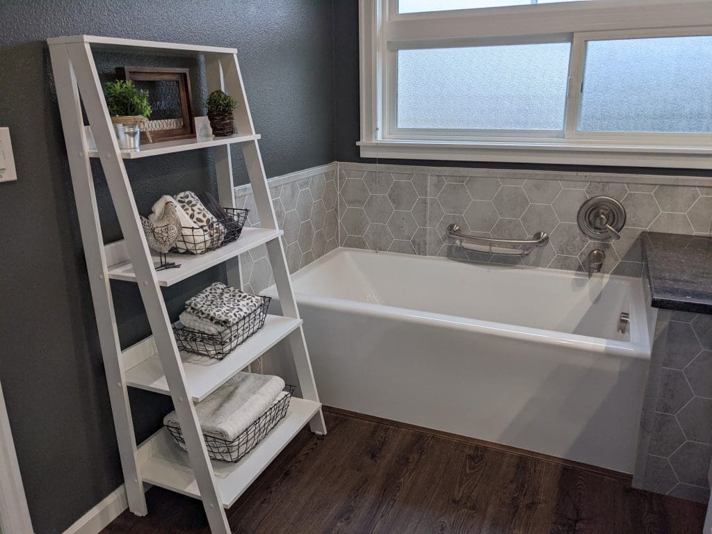 White bathtub with gray wall surround, window, and leaning storage shelf
